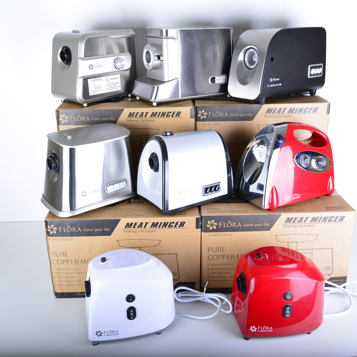 Four countertop electric meat mincers stacked on top of their original cardboard packaging boxes, with two additional mincers in white and red placed in front on a white surface against a white background.
