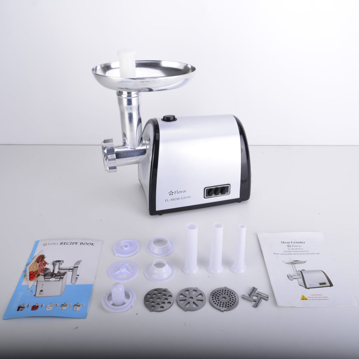A countertop meat grinder with a white and black body and silver metal hopper, shown on a light gray surface with accessories and two instruction manuals laid out in front.
