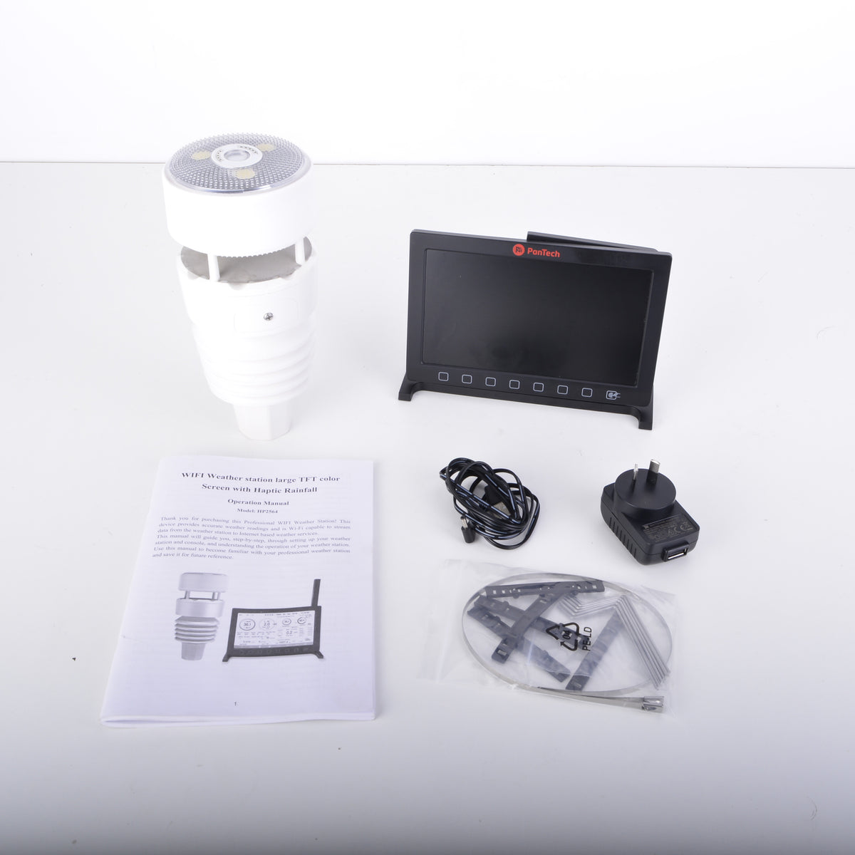 A wireless security camera kit laid out on a white surface, including a white cylindrical camera body, a small black display screen, a power adapter, cables, mounting hardware in a plastic bag, and an instruction manual.