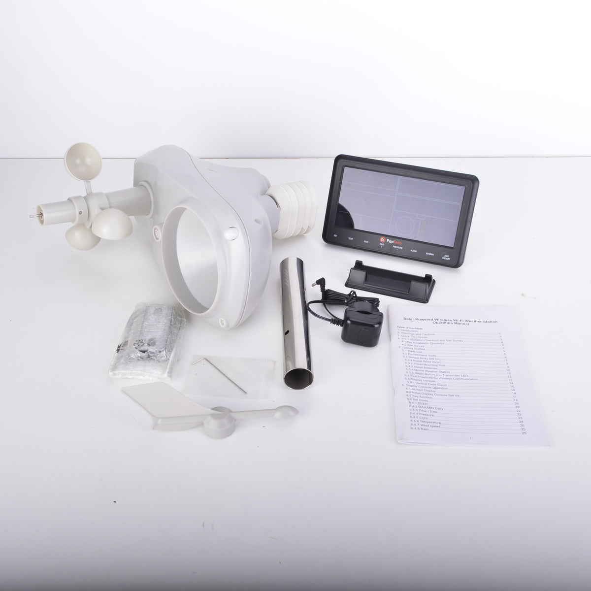 An outdoor wind speed and temperature sensor kit laid out on a white surface, including a white plastic face mask with wind cups, a small digital display screen, a black battery or charger, a metal rod, mounting hardware, a wrapped accessory, and a printed instruction sheet.